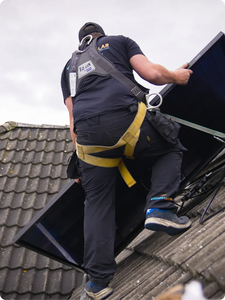 Monteur met veiligheidsharnas draagt een zwart zonnepaneel over een pannendak